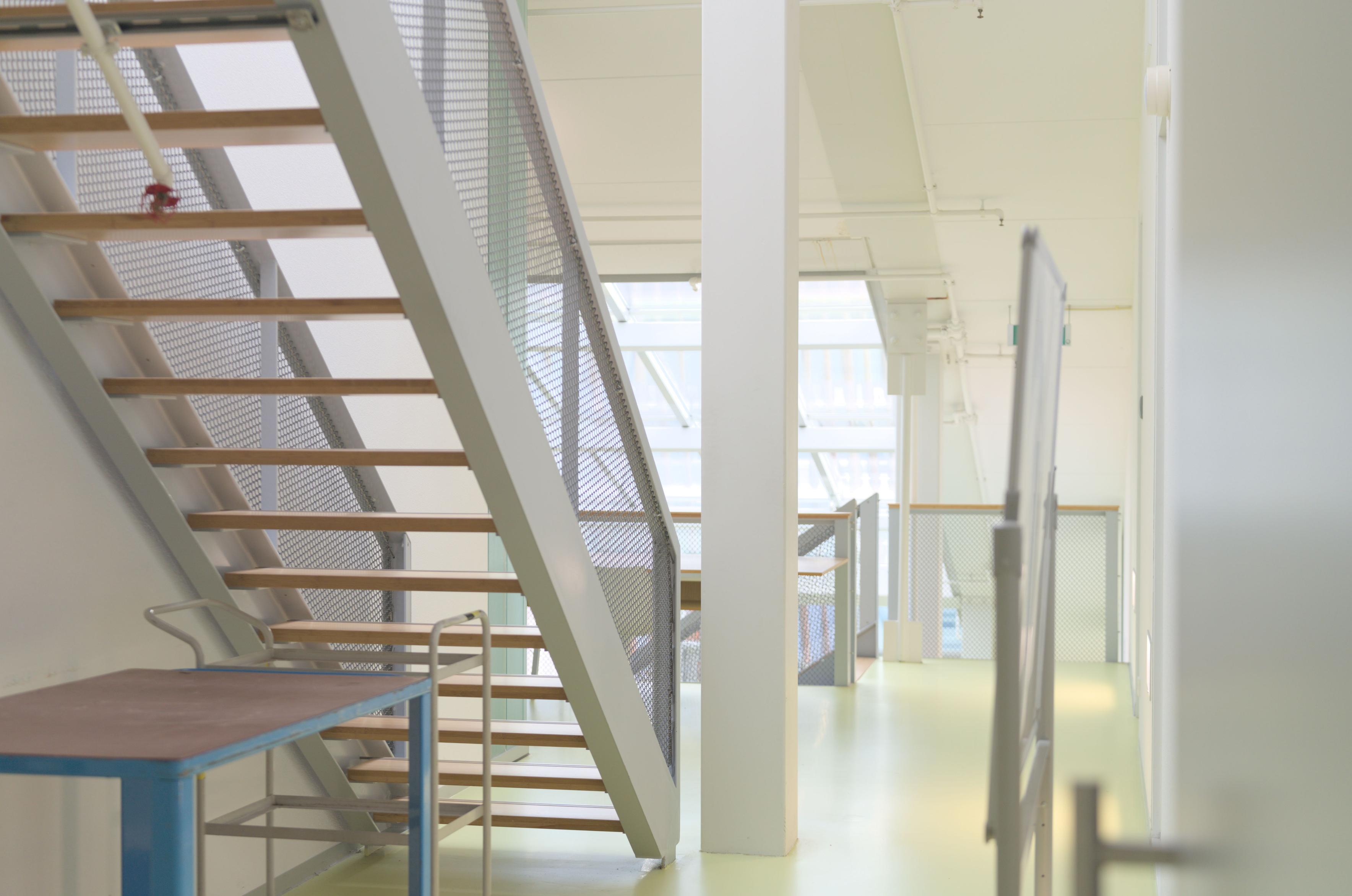 A corridor with a pastel green floor and a staircase with wooden steps. All walls are light grey. A steel beam stands vertically in the middle of the frame and in the background we see a skylight in the sloped roof of the building. In the foreground are a blue cart and a whiteboard.